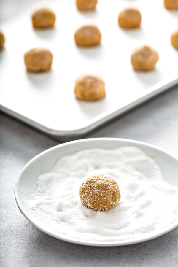 Cookie dough rolled into a ball being coated in granulated sugar with a baking sheet filled with cookie dough balls in the background.