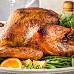 A roasted Thanksgiving turkey with golden brown and crispy skin on a serving platter with a bowl of cranberry sauce in the background.