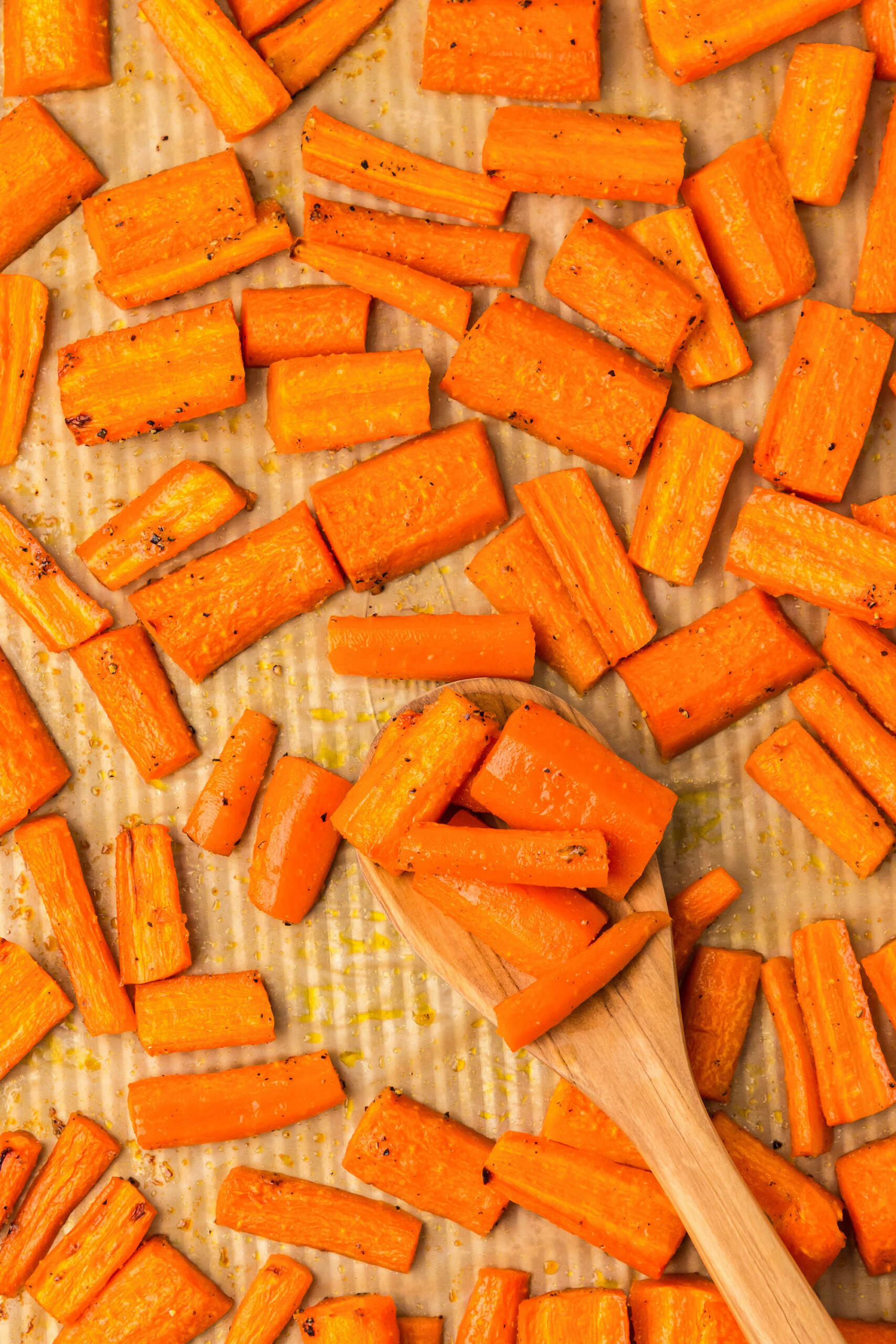 Honey glazed carrots on a baking sheet lined with parchment paper with a wooden spoon scooping up a serving of roasted carrots.