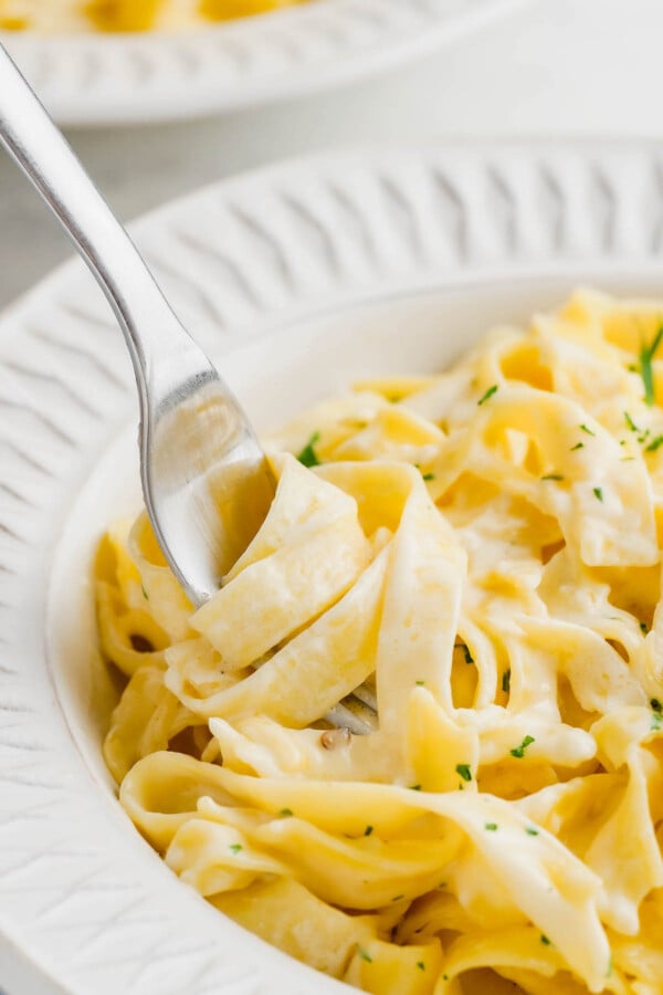 A fork scooping up a bite of fettuccini alfredo off of a plate.