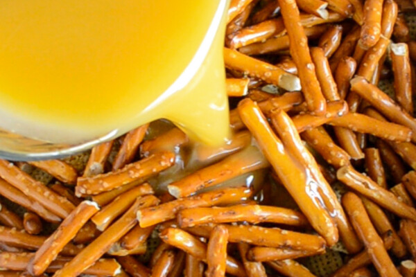 Butter mixture being poured over the top of a bowl of pretzels and Chex cereal.
