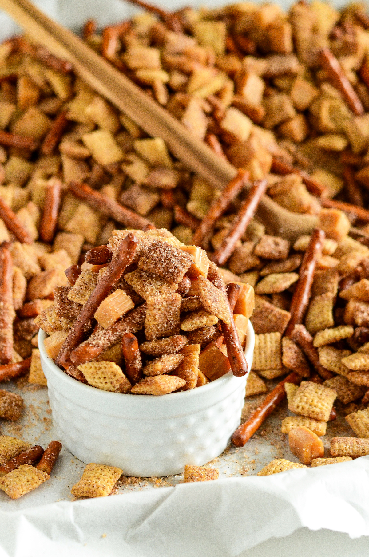 A bowl of cinnamon churro Chex mix set on a cookie sheet filled with the snack mix with a wooden spoon for serving.