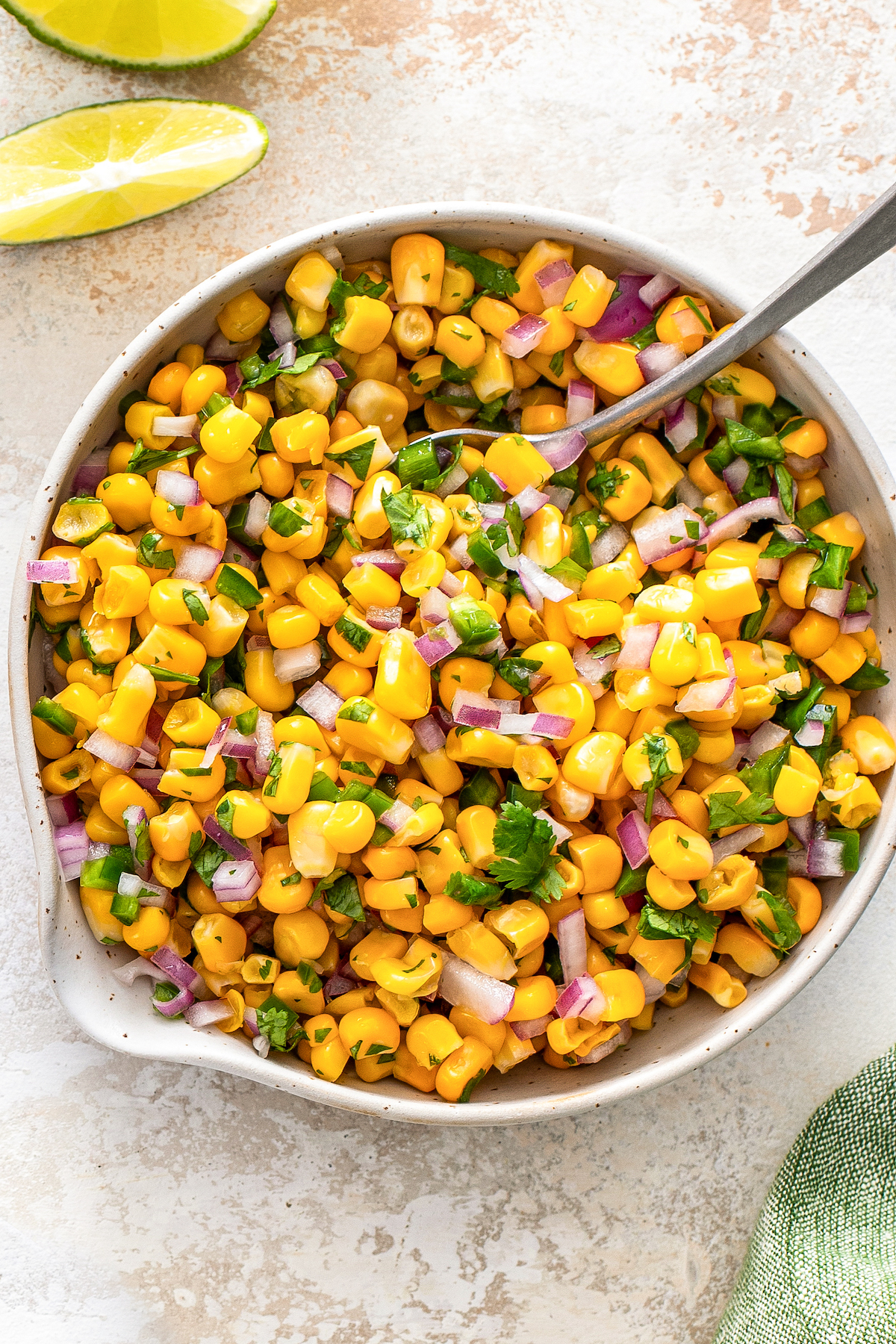 Refreshing corn salsa with jalapeño, red onion and cilantro in a bowl with a spoon for serving and lime wedges on the side. 