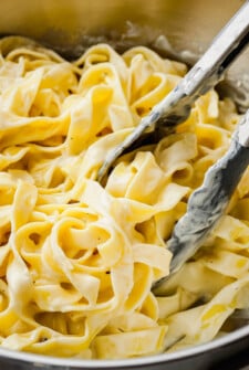 Tongs scooping up a serving of pasta coated in a rich and creamy homemade alfredo sauce out of a sauce pot.