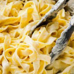 Tongs scooping up a serving of pasta coated in a rich and creamy homemade alfredo sauce out of a sauce pot.