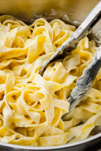 Tongs scooping up a serving of pasta coated in a rich and creamy homemade alfredo sauce out of a sauce pot.