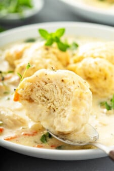 A bowl of homemade chicken and dumplings with a spoon holding a homemade dumpling with a bite taken out of it to show the fluffy center.