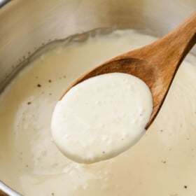 A wooden spoon scooping out a serving of a rich and creamy homemade alfredo sauce out of a pot.