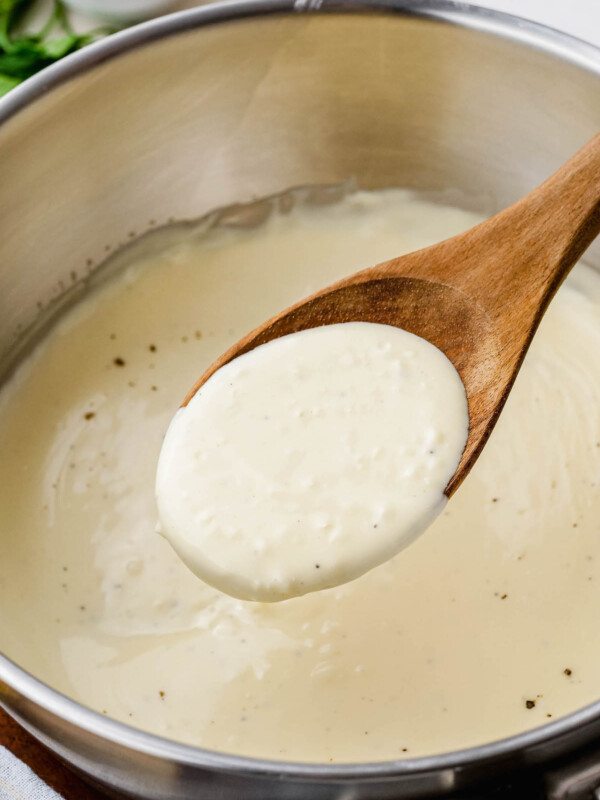 A wooden spoon scooping out a serving of a rich and creamy homemade alfredo sauce out of a pot.
