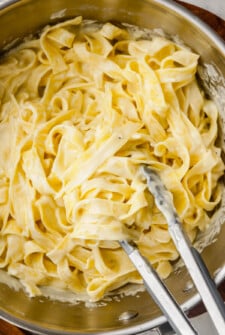 Homemade Alfredo sauce being tossed with cooked fettuccine noodles with tongs in a sauce pot.