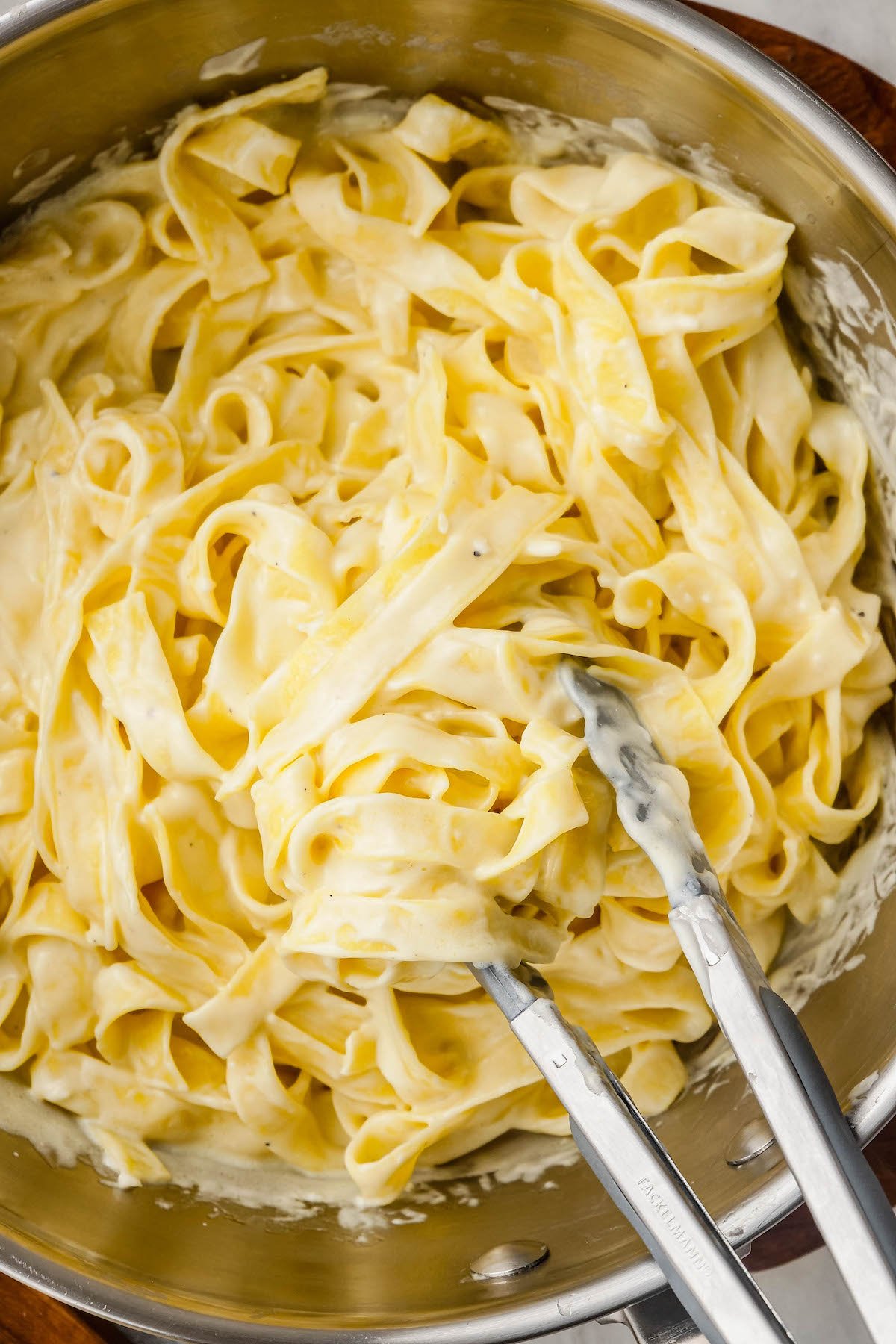 Homemade Alfredo sauce being tossed with cooked fettuccine noodles with tongs in a sauce pot.