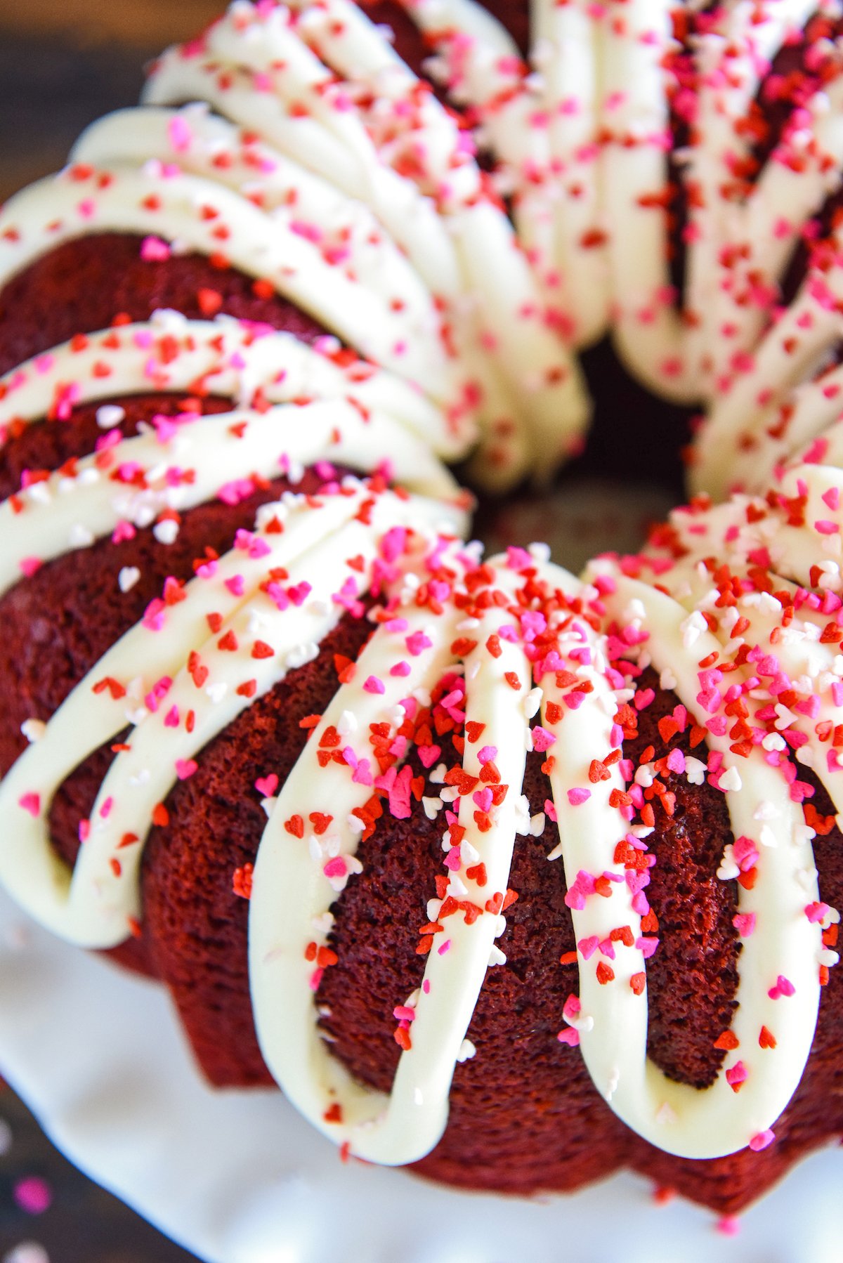 Red, white and pink sprinkles sprinkled on top of cream cheese icing drizzled on top of a red velvet bundt cake on a cake stand.