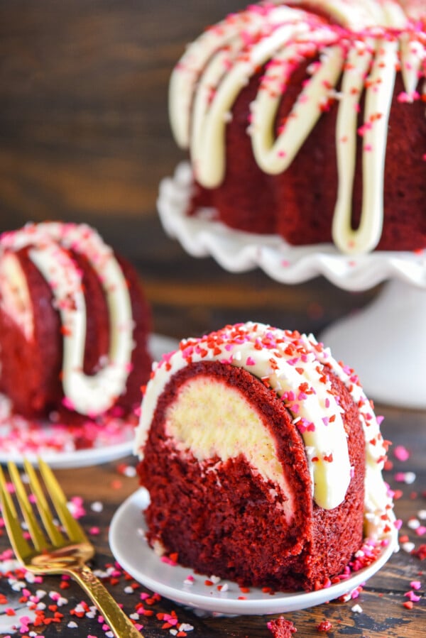 Two slices of red velvet bundt cake on plates with the full bundt cake on a cake stand in the background.