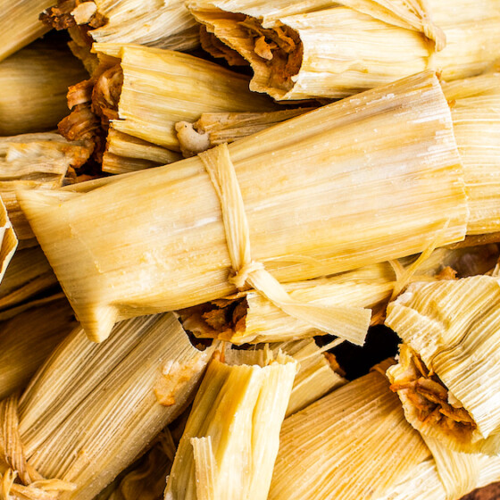 Pork tamales wrapped in corn husks stacked on top of each other on a wooden board.