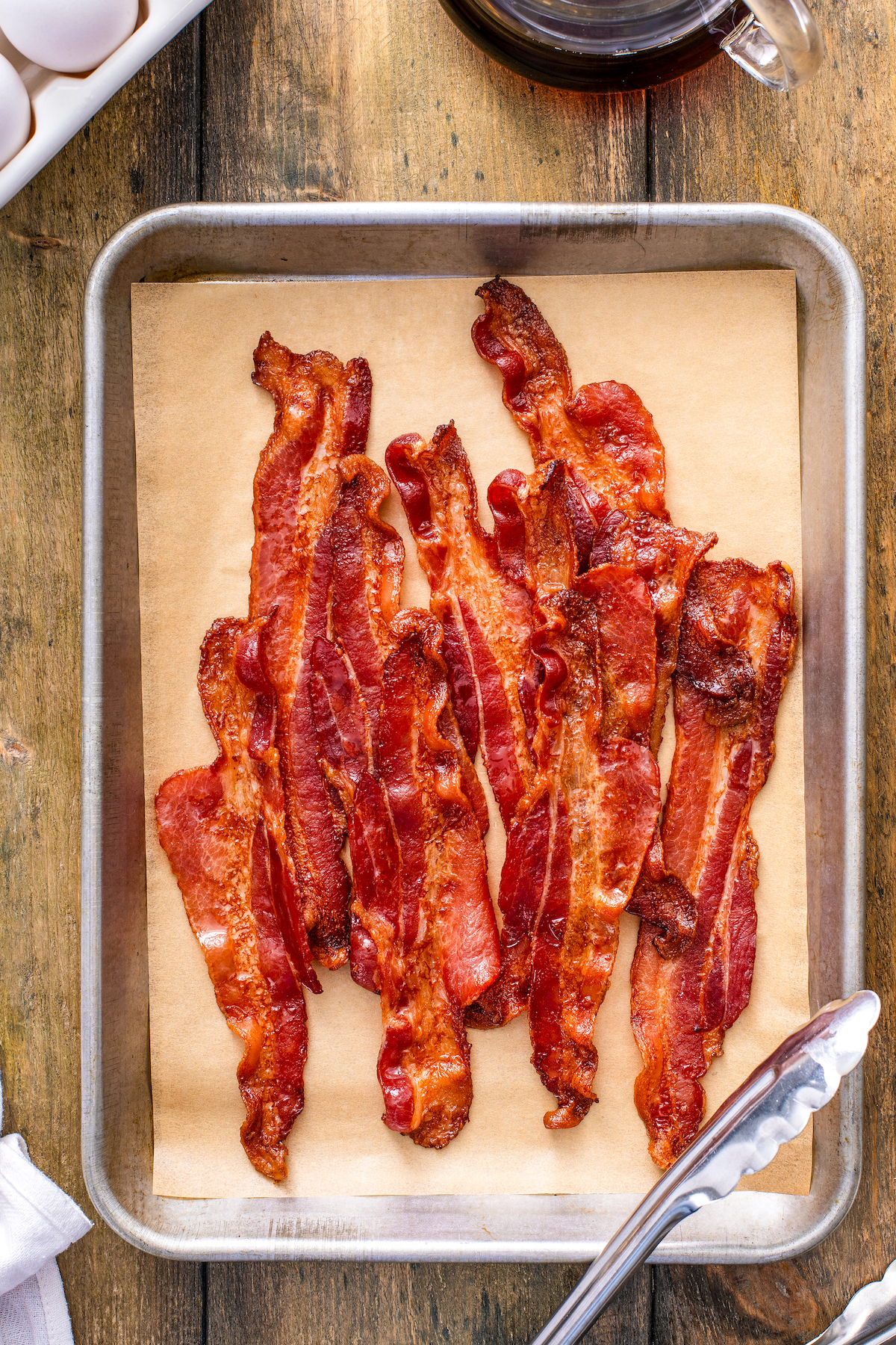 Crispy cooked bacon in oven after draining on paper towels. Bacon is arranged on top of a parchment paper lined baking sheet with tongs.