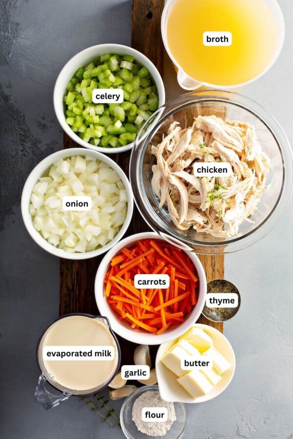 Ingredients for chicken and dumplings recipe arranged in bowls on a cutting board, from top to bottom: broth, celery, chicken, onion, carrots, thyme, evaporated milk, garlic, butter and flour.