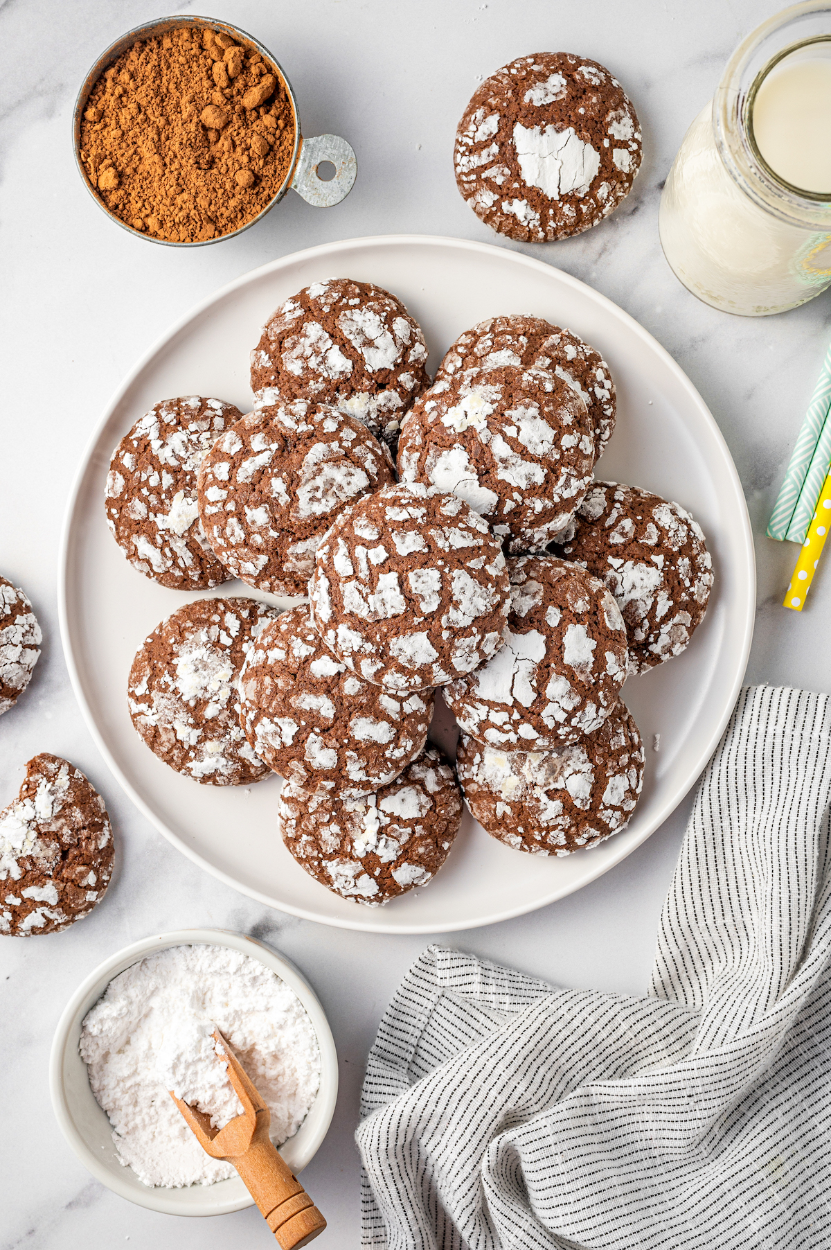 A platter of homemade chocolate crinkle cookies coated in powdered sugar on a serving plate with a glass of milk.