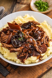 Crock pot pot roast with a rich brown gravy served over a bed of mashed potatoes on a plate.