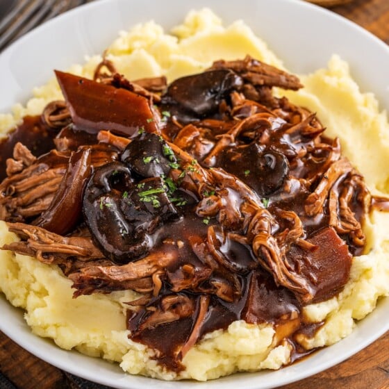 Crock pot pot roast with a rich brown gravy served over a bed of mashed potatoes on a plate.