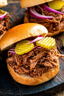 Pulled pork sandwiches on a wooden serving board with pickles and red onions on top.
