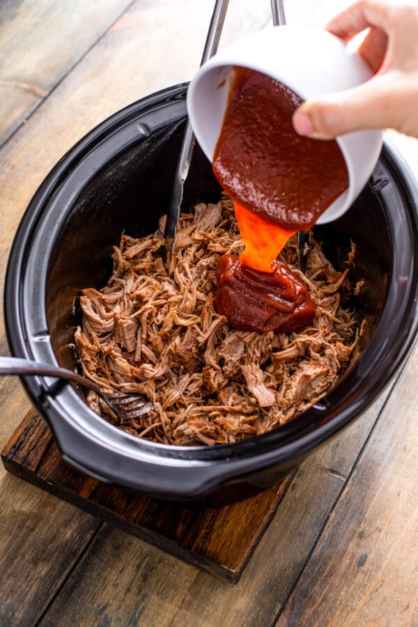 Pulled pork in crock pot with two forks and bbq sauce being poured over the top of the shredded pork.
