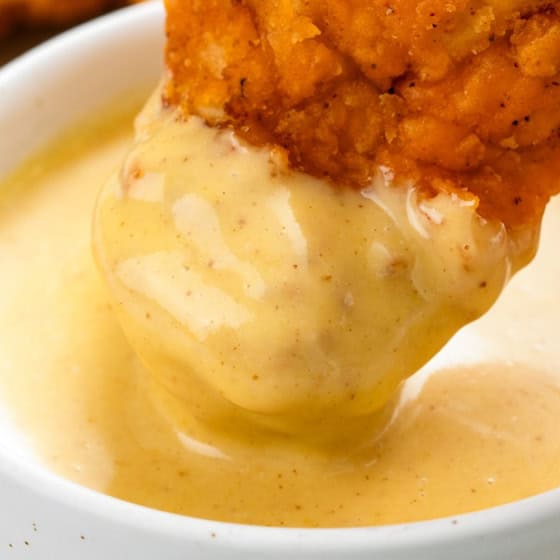 A golden chicken tender is dipped into smooth honey mustard.