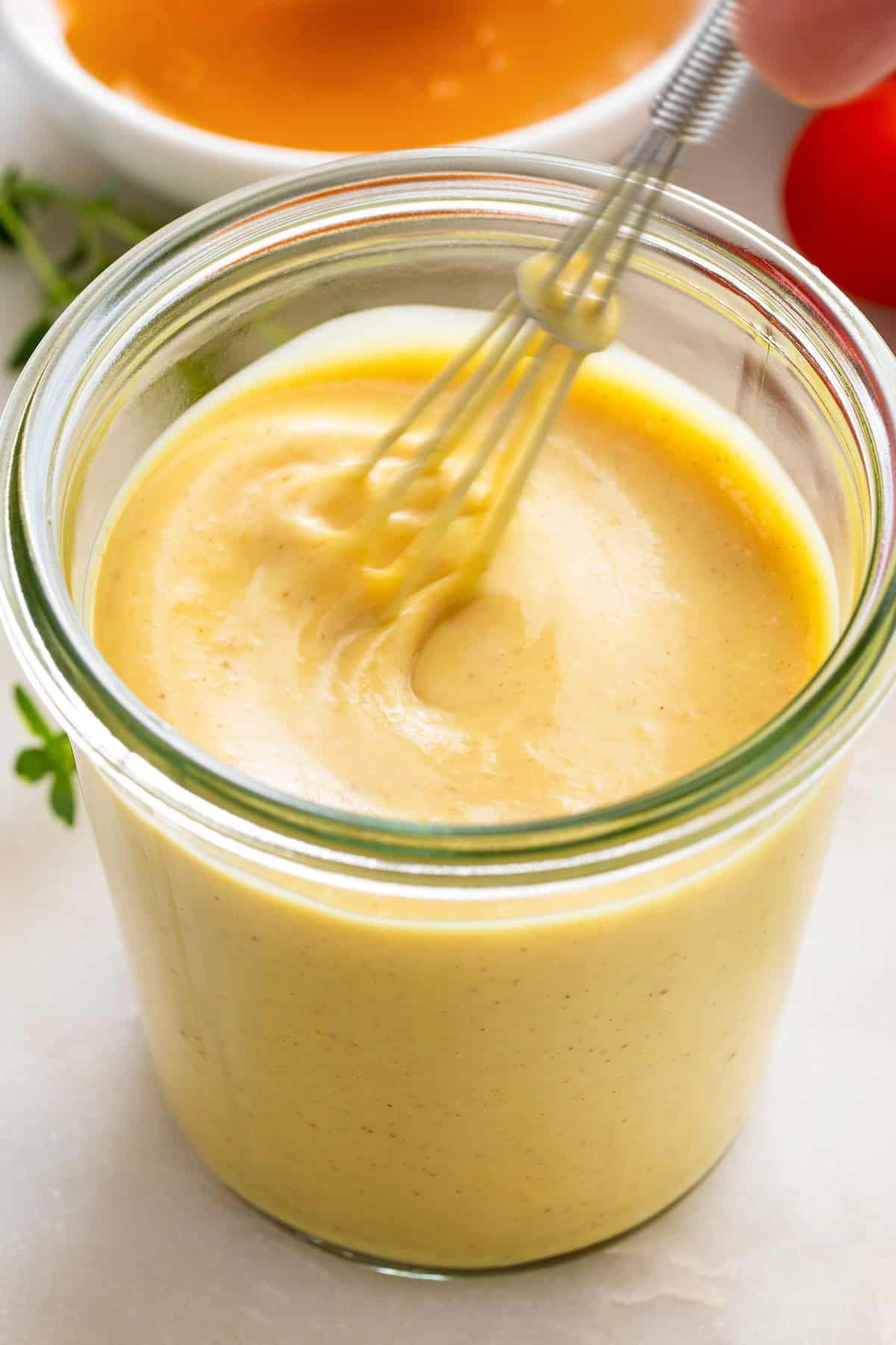 A whisk is stirring honey & mustard with other ingredients together in a jar.