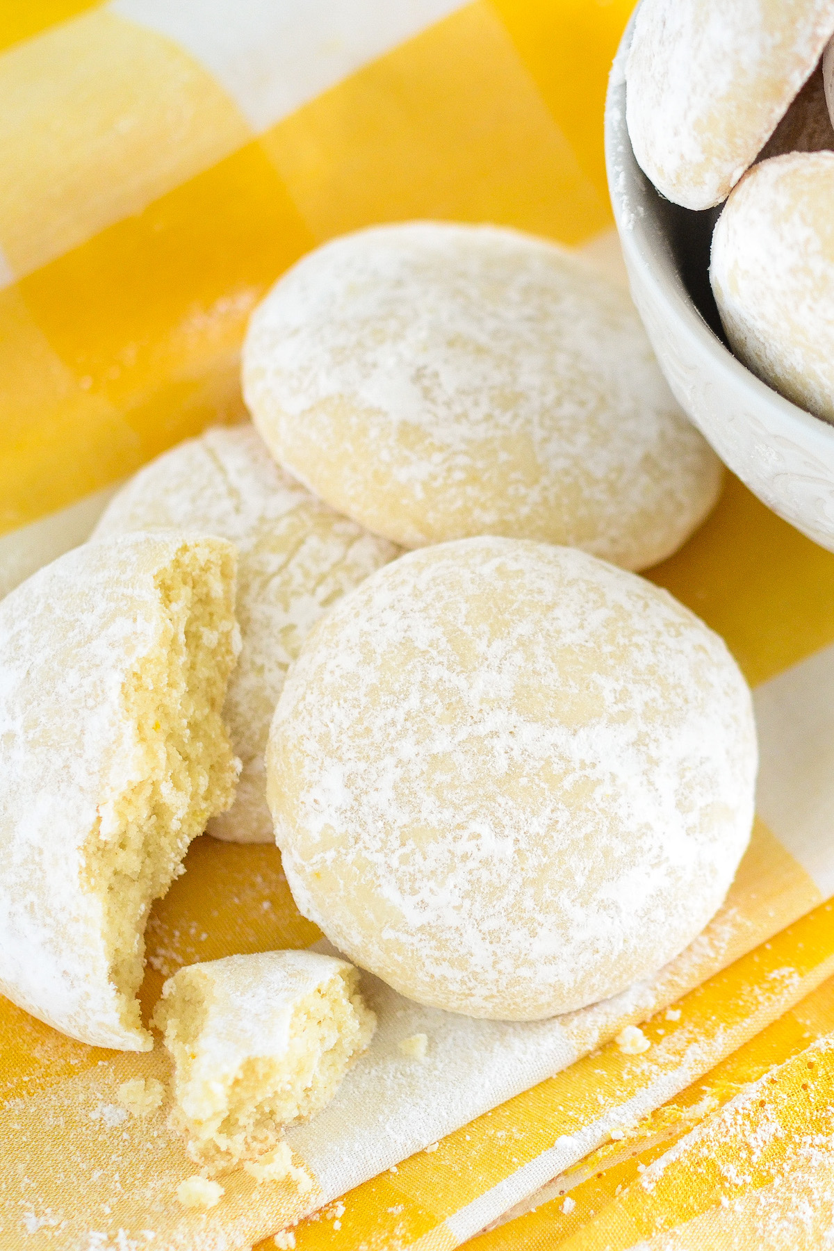 Lemon cooler cookies on a yellow napkin with a bowl of cookies in the background and one cookie with a bite taken out of it.