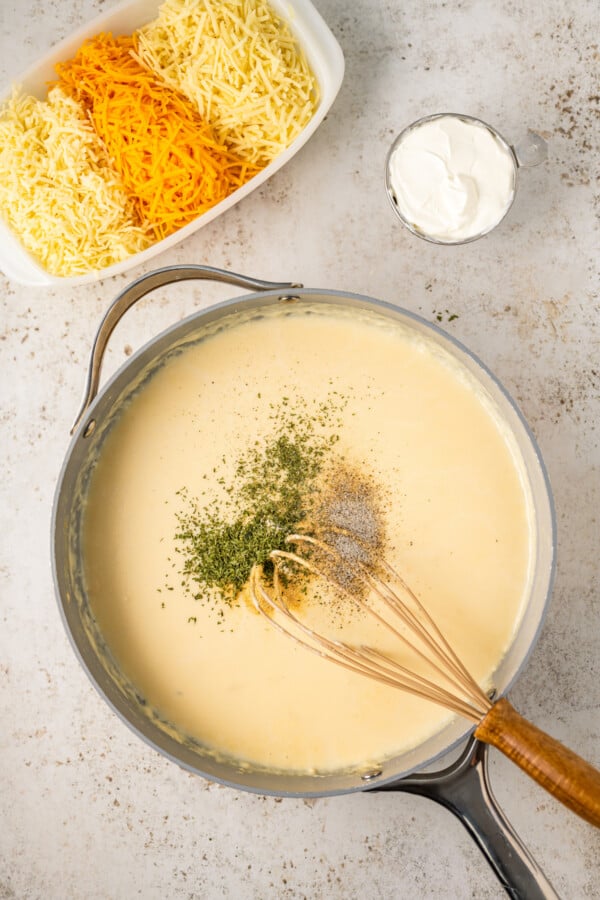 A sauce pan filled with creamy homemade cheese sauce with seasonings being added and whisked in.