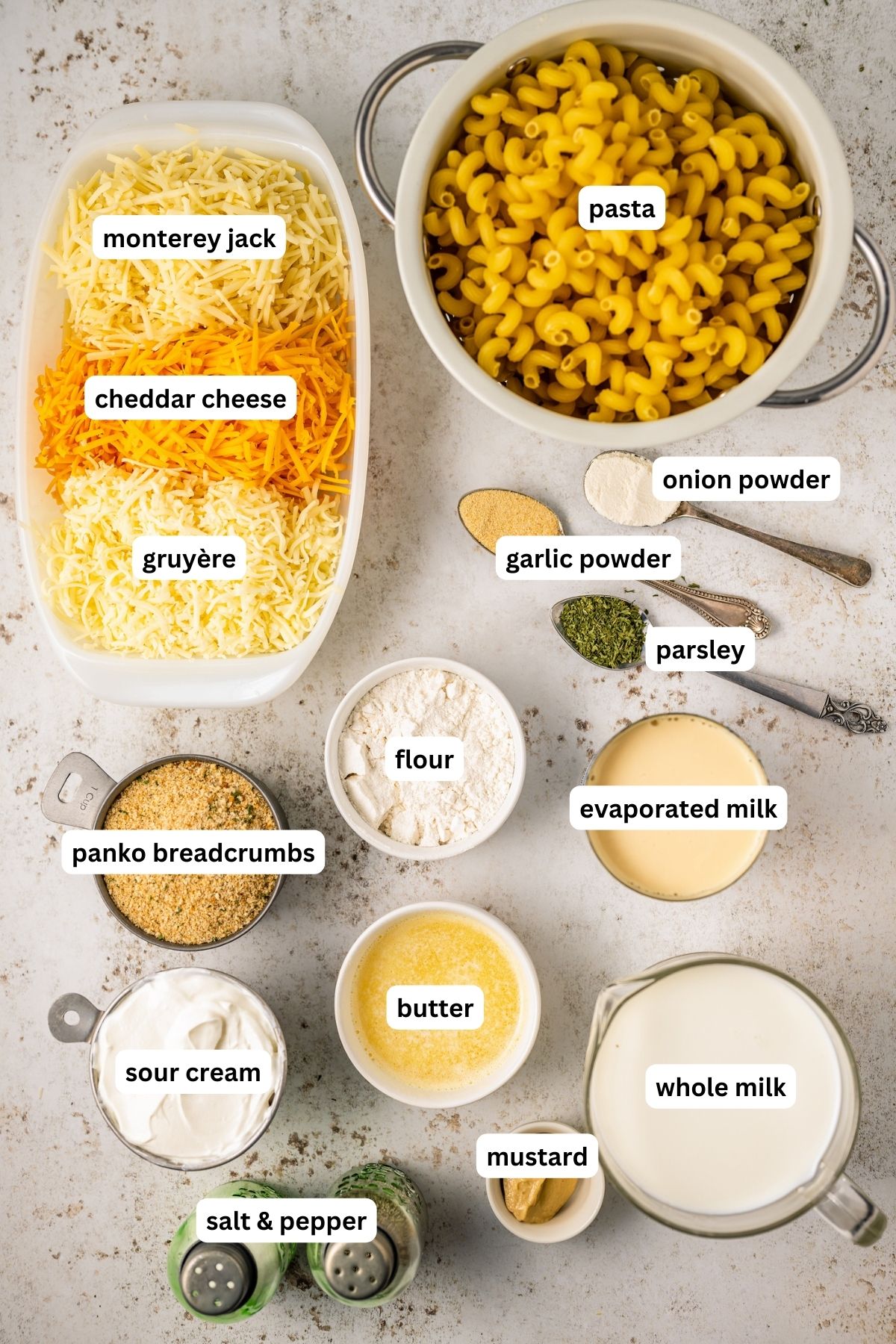 Ingredients in bowls for mac and cheese recipe, arranged from top to bottom: pasta, Monterrey jack cheese, cheddar cheese, gruyere cheese, onion powder, garlic powder, dried parsley, flour, Panko Breadcrumbs, evaporated milk, sour cream, butter, whole milk, mustard, salt and pepper.