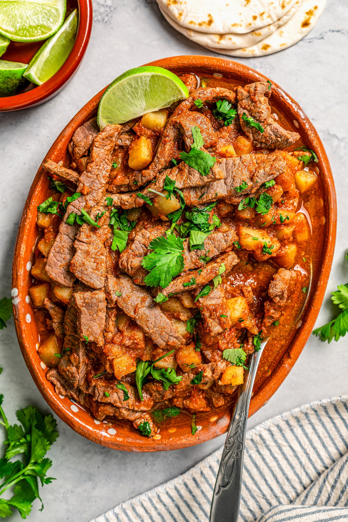 Juicy and spicy Bistec Ranchero with potatoes served on a large serving platter with fresh cilantro on top and lime wedges on the side.