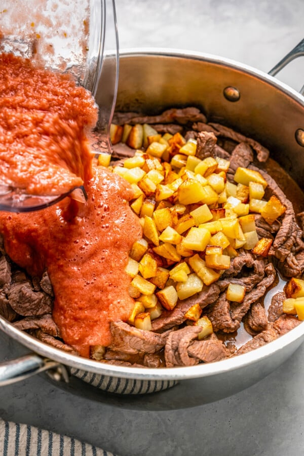 Ranchero sauce being poured into a skillet with seared strips of beef and browned potatoes.