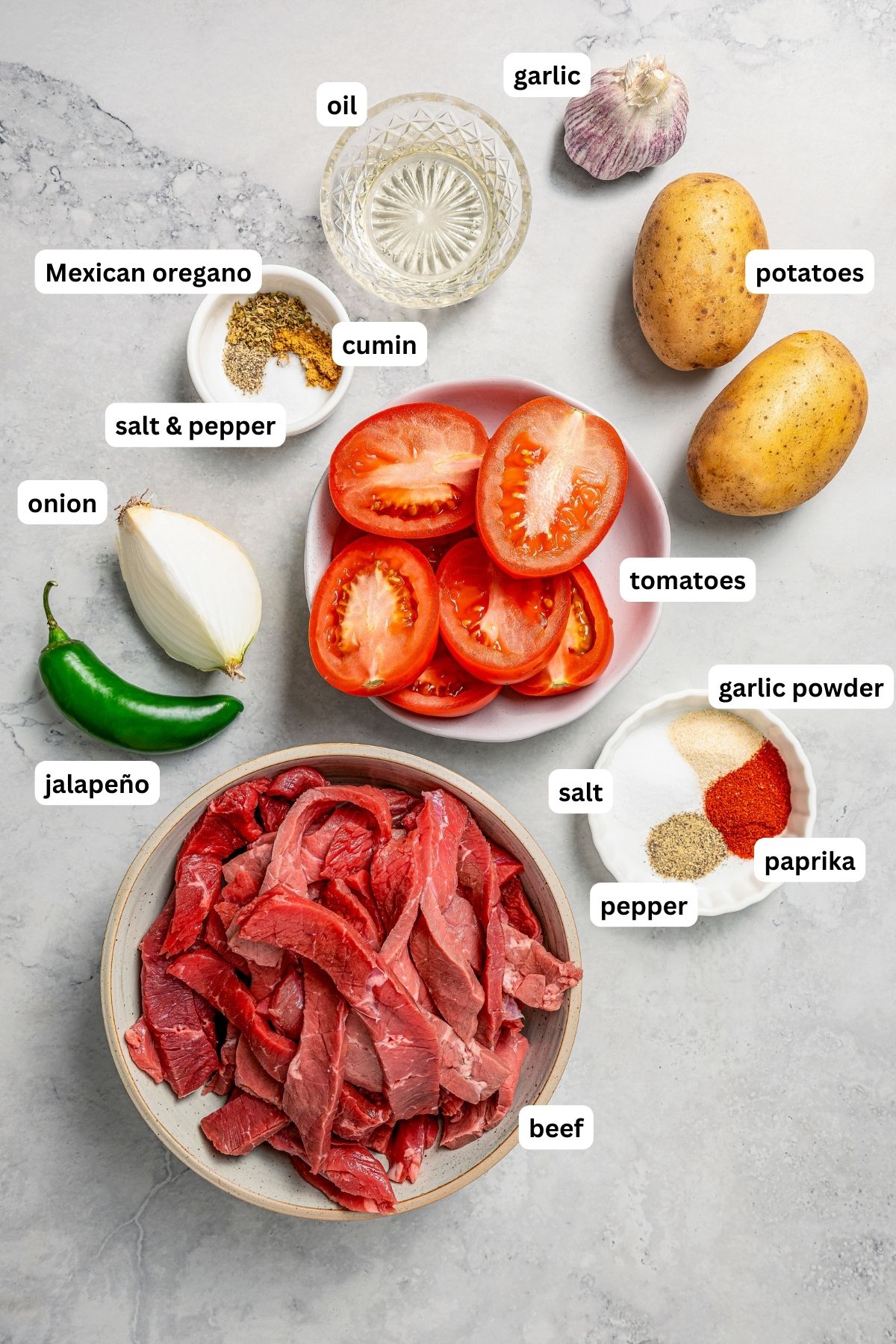 Ingredients for bistec ranchero recipe, from top to bottom: garlic, oil, potatoes, mexican oregano, cumin, salt, pepper, tomatoes, onion, jalapeño, garlic powder, paprika and sliced beef.