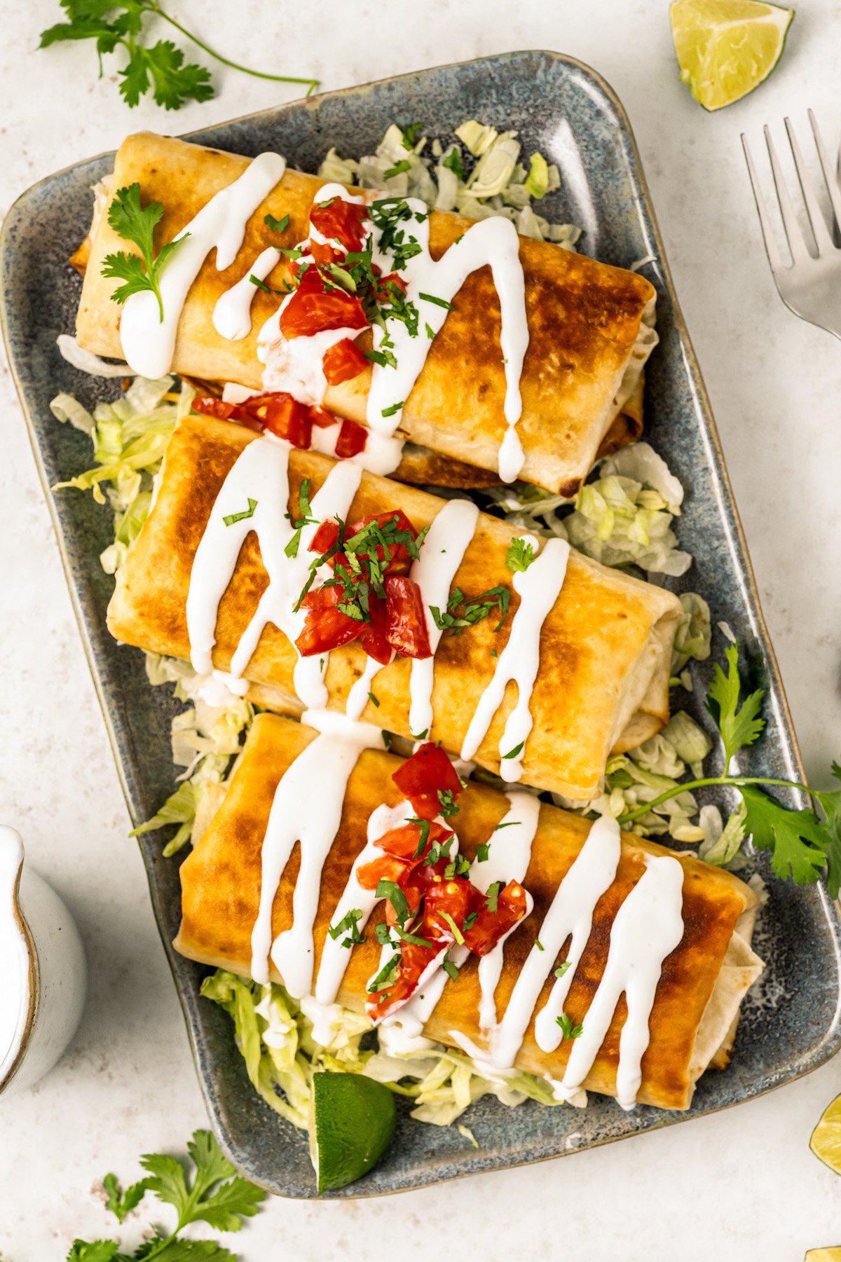 Three crispy chimichangas on a serving plate topped with pico de gallo, lettuce, and Mexican crema.