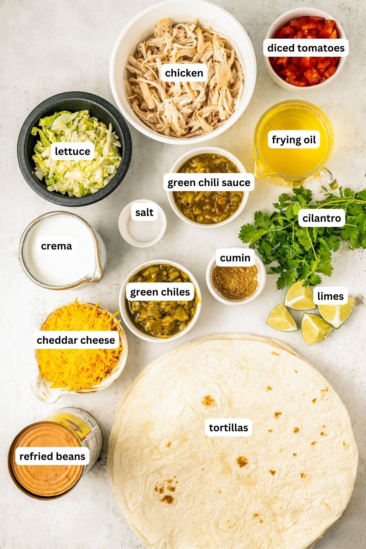 Ingredients for chicken chimichanga recipe, arranged in bowls. From top to bottom: diced tomatoes, shredded chicken, lettuce, frying oil, green chili sauce, salt, Mexican Crema, cilantro, cumin, green chilies, cheddar cheese, limes, refried beans and large tortillas.