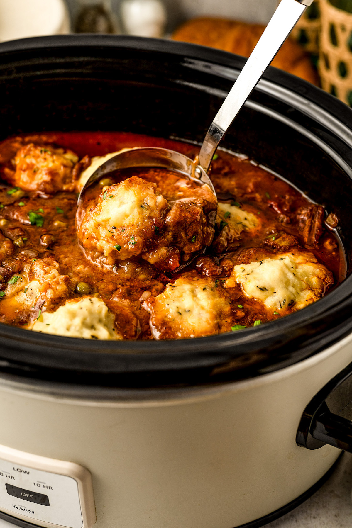 A ladle scooping out a serving of beef stew with dumplings from a crockpot.