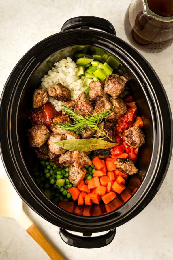 Filling the crockpot with seared beef, vegetables, herbs and beef broth.