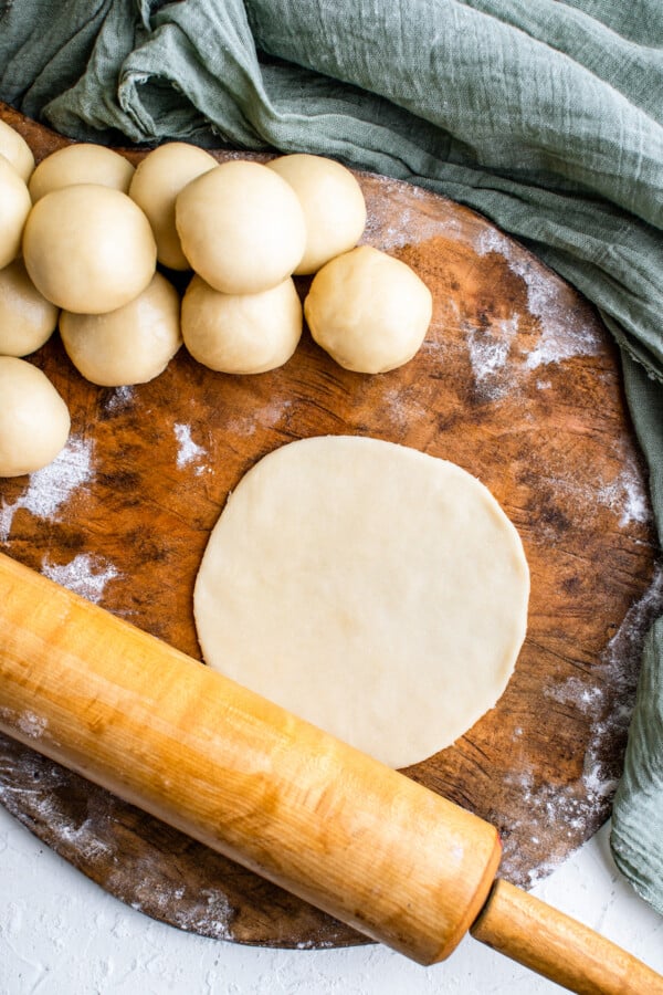 Balls of empanada dough stacked on the side with a rolling pin rolling a dough ball flat into a round disk.