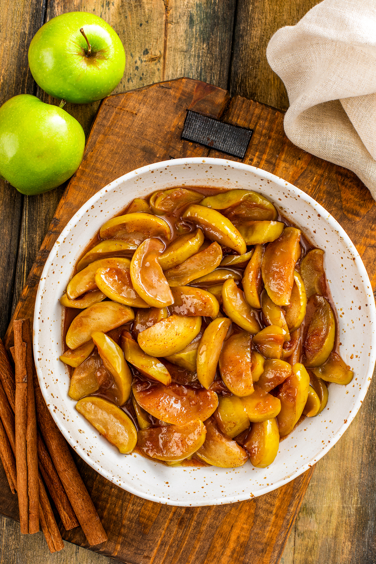 Overhead view of a bowl of fried apples with cinnamon sticks and two green apples on the side of the bowl.