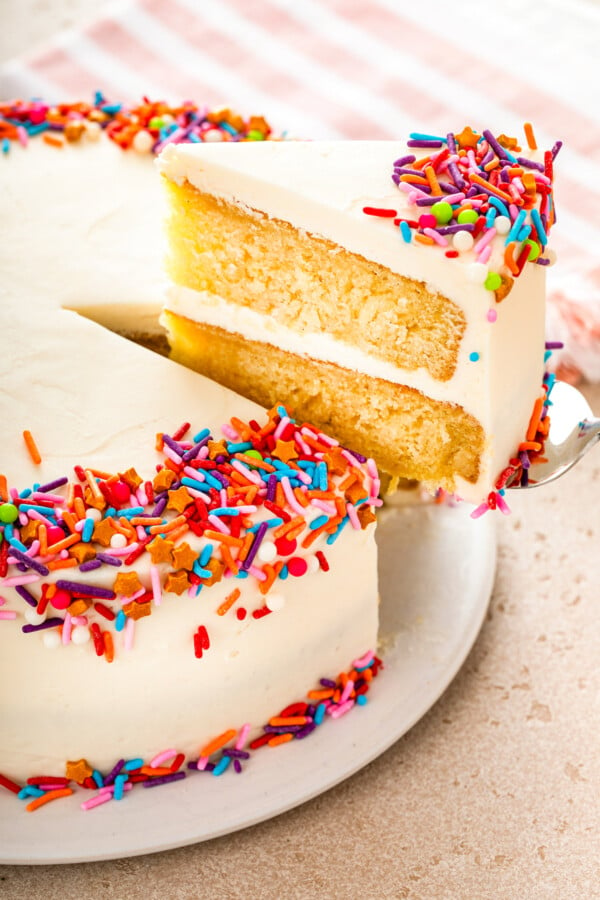 A slice of moist vanilla cake layered with buttercream frosting being lifted with a spatula from the whole cake on a cake plate.