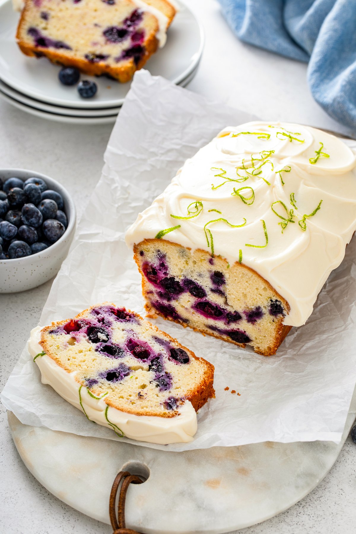 Lime Blueberry Pound Cake topped with creamy frosting and fresh lime zest being sliced into pieces on a parchment paper lined cutting board.