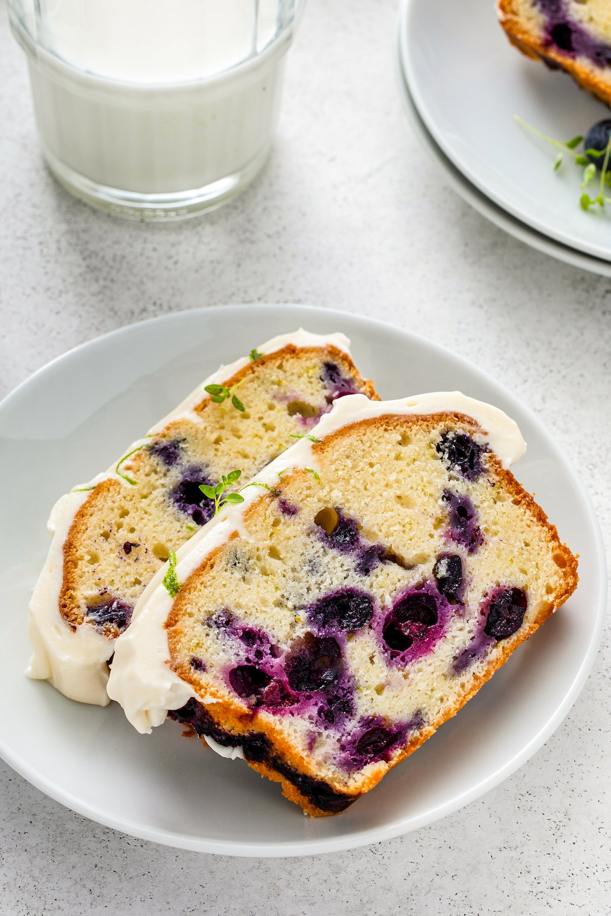 Moist and dense cream cheese pound cake loaded with blueberries and lime zest and topped with cream cheese frosting sliced into pieces and served on a plate.