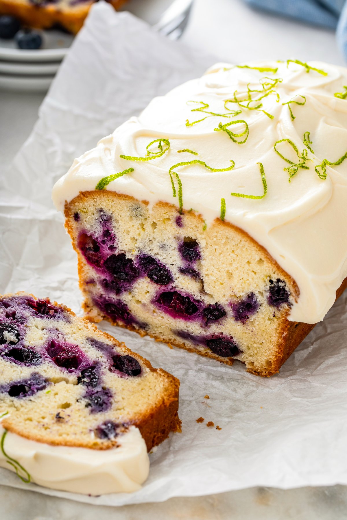 A sweet and dense lime blueberry pound cake topped with a thick layer of cream cheese frosting being sliced into pieces.