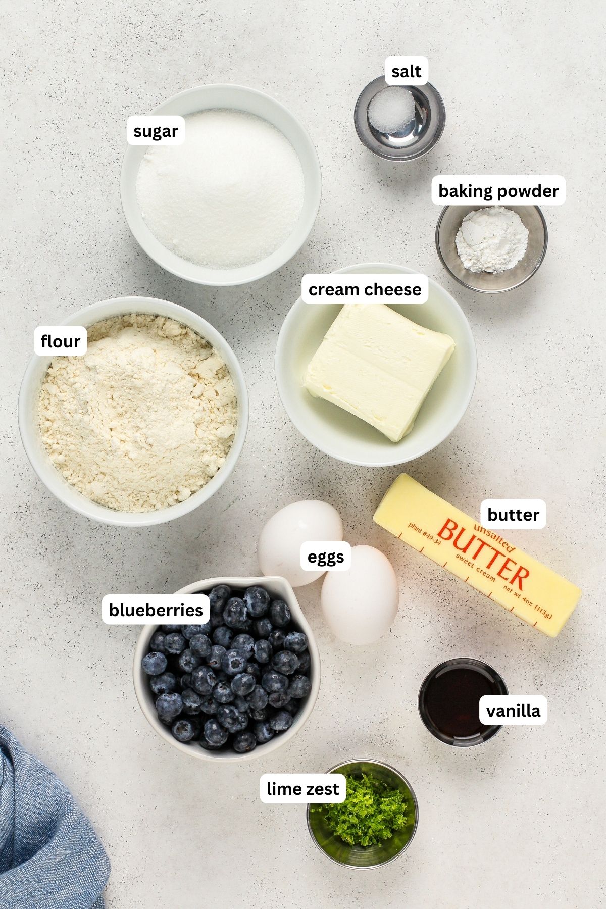 Ingredients for blueberry pound cake recipe arranged in bowls, from top to bottom: salt, sugar, baking powder, cream cheese, flour, butter, eggs, blueberries, vanilla and lime zest.