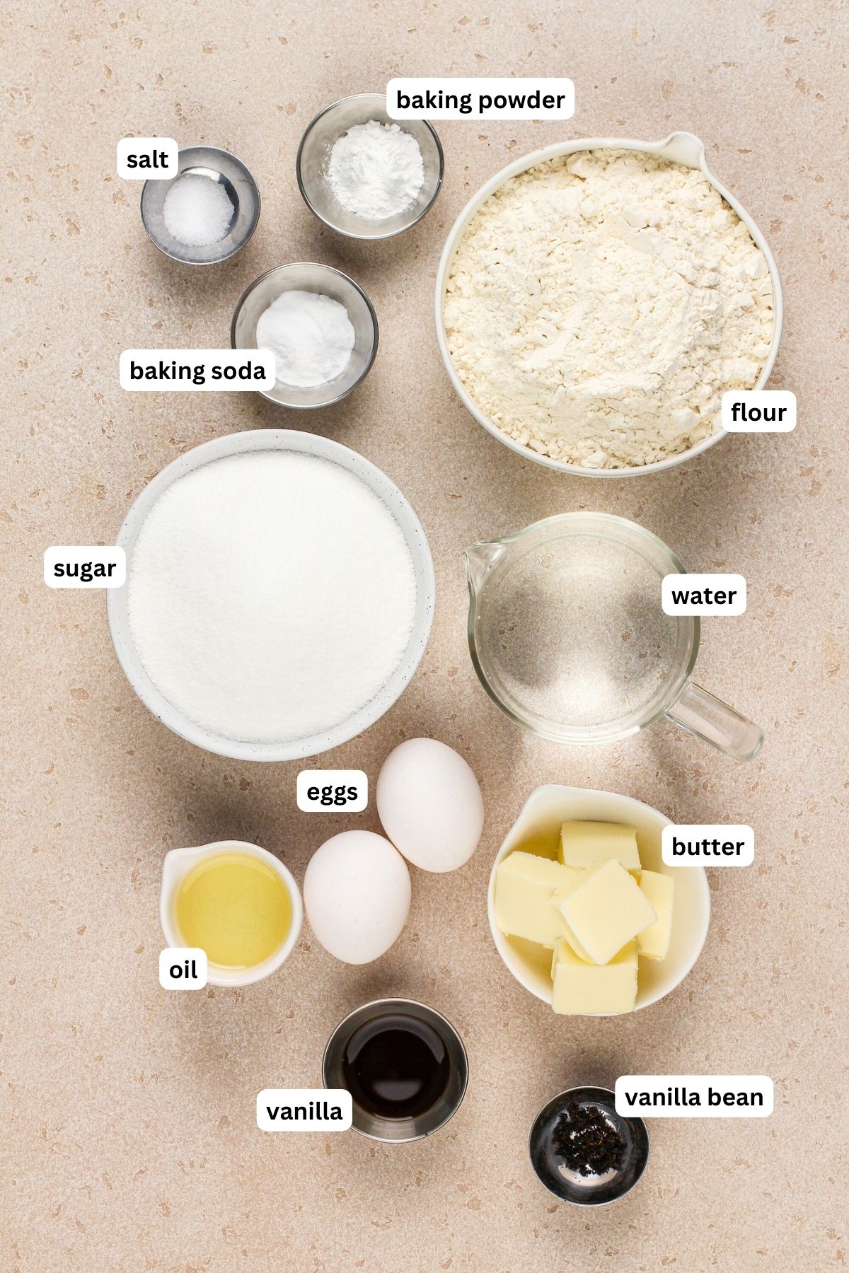 Ingredients for vanilla cake recipe arranged in bowls, from top to bottom: baking powder, salt, baking soda, flour, granulated sugar, water, eggs, oil, butter, vanilla extract and vanilla bean.