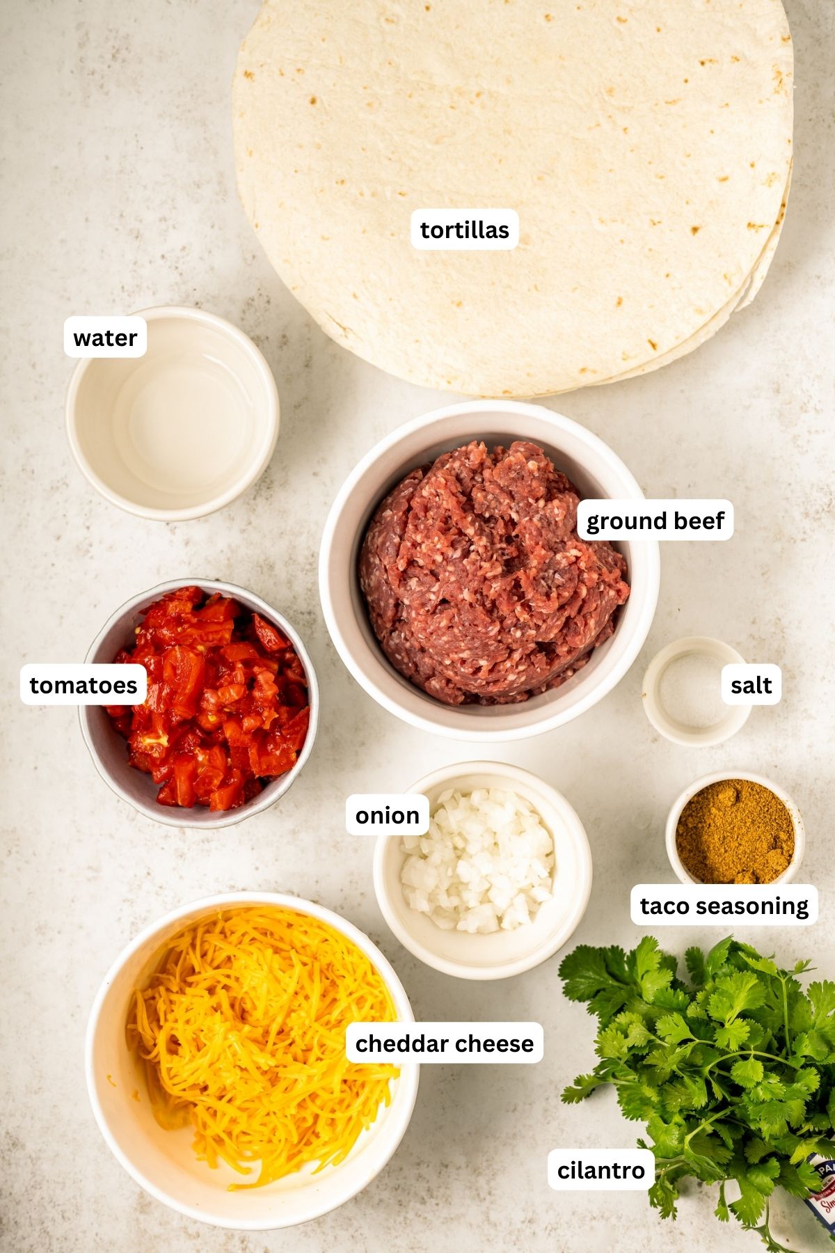 Ingredients arranged in bowls for Taco Bell Meximelt recipe, from top to bottom: tortillas, water, ground beef, tomatoes, salt, onion, taco seasoning, cheddar cheese and cilantro.