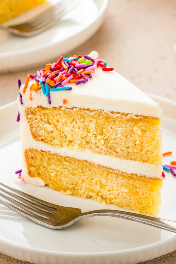 A slice of moist vanilla cake layered with fluffy vanilla buttercream with rainbow sprinkles on a plate.