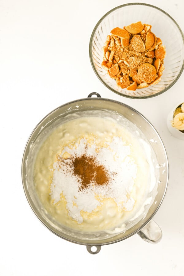 Creamy whipped mixture in a bowl with cinnamon and instant banana pudding mixture being added.