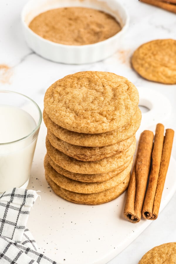 Chewy snickerdoodles stacked on top of each other with cinnamon sticks and a glass of milk on the side.