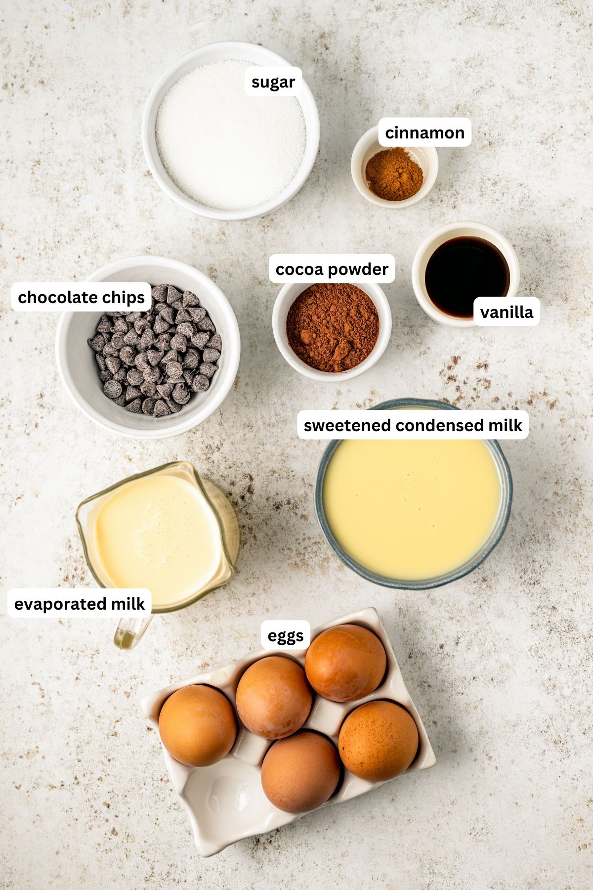 Ingredients for chocolate flan recipe arranged in bowls, from top to bottom: granulated sugar, ground cinnamon, chocolate chips, cocoa powder, vanilla extract, sweetened condensed milk, evaporated milk and eggs. 